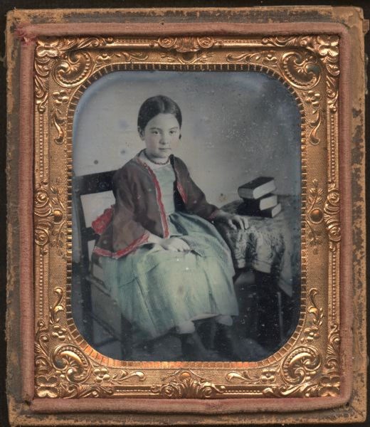Sixth plate ferrotype/tintype of unidentified young girl. She is sitting on a chair, with her left hand resting on a cloth-covered table on which three books are stacked, and her right hand in her lap. She is wearing an open jacket with bell sleeves over a striped pinafore, and a hand-colored red necklace. She appears to be sitting on a book. The jacket has a hand-colored red border, and the dress is blue. Her hair is pulled back and her cheeks hand-colored. 