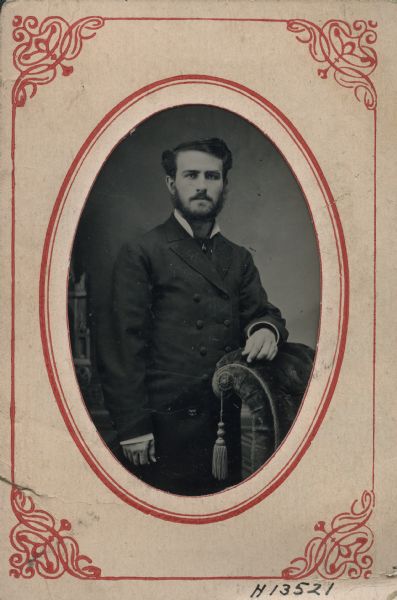 Sixth plate ferrotype/tintype. Three-quarter length portrait of Doctor John M. Evans, from Evansville, Wisconsin, standing in front of a painted backdrop. He is wearing a suit, necktie with pin, and has a beard and moustache. He has one arm resting on the curved back of an upholstered chair.