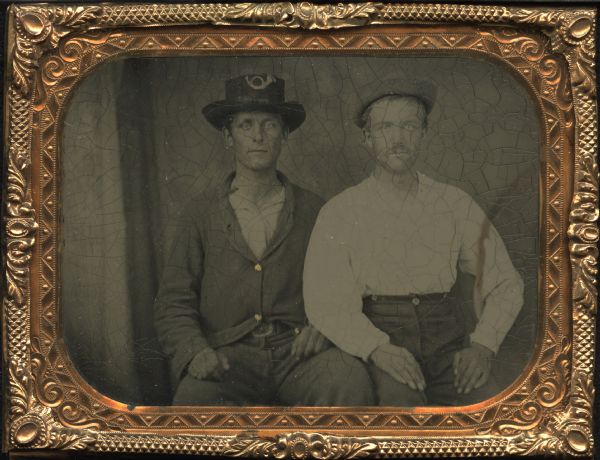 Quarter plate ferrotype/tintype of two unidentified men. Three-quarter length portrait of the men sitting, facing forward, with their hands on their laps. The man on the left is wearing a buttoned coat and brimmed hat with bugle ornament on crown. The man on the right also wears a hat, and is simply dressed in a white shirt and trousers. Gold details on buttons. 