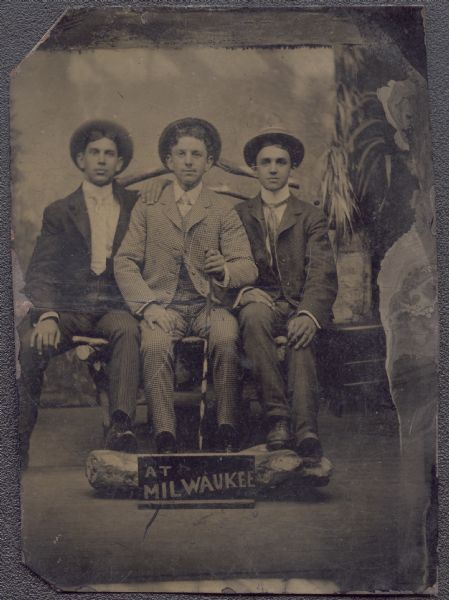 Ferrotype/tintype full-length portrait of three young unidentified men wearing hats, sitting on a twig bench in front of a painted backdrop, facing forward. They have their feet perched on a log, with a painted sign on the floor in front that reads" "At Milwaukee." Each man is wearing a suit with a necktie. The man in the center is wearing a checked suit with the collar folded down, and is holding a cane. The men on either side have stand-up collars. 