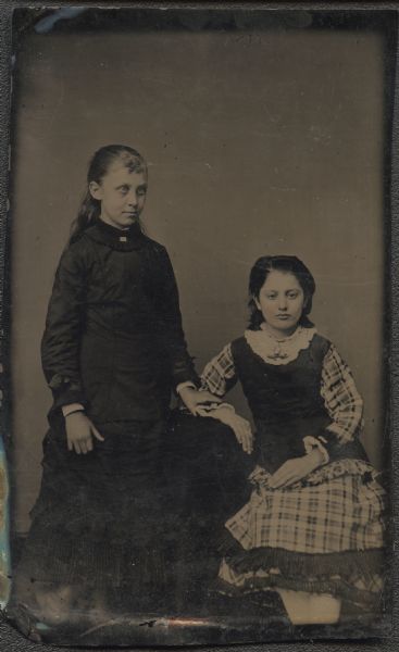 Ferrotype/tintype of Louisa Zimmer and Ada (?) Empey. The girl on the left is standing, facing halfway right, wearing a dark dress and ruched collar, her hand resting on the elbow of the other girl. The girl on the right is sitting, facing front, with her elbow resting on a table. She is wearing a plaid skirt and matching plaid sleeves with a dark bodice, and a white lace collar with brooch. Cheeks are hand-colored. 