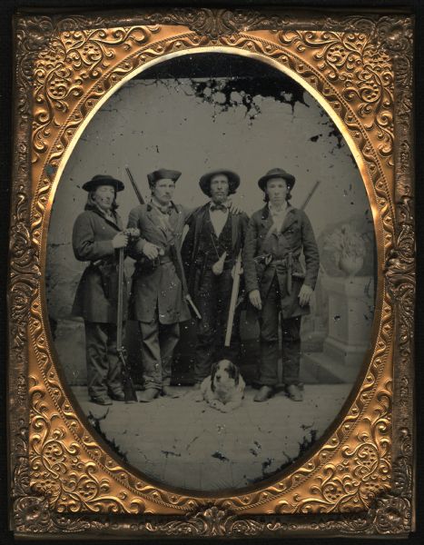 Half plate ambrotype of (left to right) Ludwig Boehmer, Christian Boehmer, Siegfried Mittelstadt, and Jacob Boehmer standing in front of a painted backdrop. They are standing side by side against a studio backdrop, with a dog laying at their feet. Each man is carrying a long gun and is wearing an overcoat and hat. They also carry powder flasks and knives at their waists. 