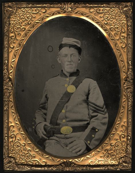 Half plate ferrotype/tintype of a three-quarter length portrait of John W. Fonda, Company C, 6th Regiment, Wisconsin Volunteer Infantry. Seated half figure facing front, left hand on left leg, right hand on cartridge box on belt. Hand-coloring on cheeks, gold details on buttons, belt buckle and medallion on sash. He is wearing the original state-issue gray uniform of the regiment. Fonda was from Prairie du Chien, and enlisted April 20, 1861.