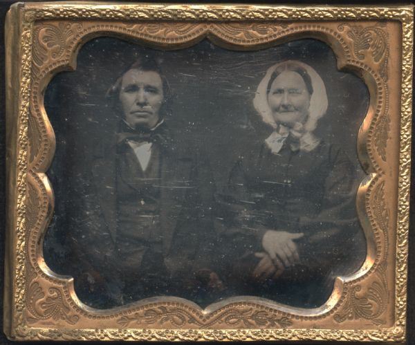 Sixth plate daguerreotype of Lamont and his wife, Scottish immigrants. Waist-up portrait, seated facing forward. He is wearing a suit and tie, and she is wearing a dark dress, white collar, and white bonnet tied under her chin, with her hands crossed on her lap. 