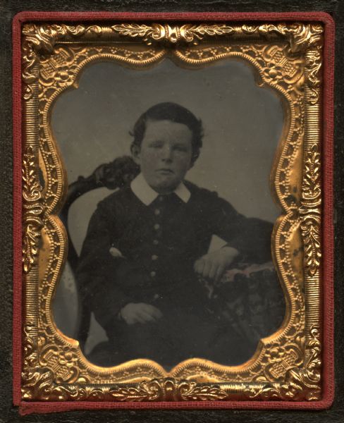Ninth plate ferrotype/tintype of Henry E. Knapp, seated on a balloon-back chair, left forearm resting on cloth-covered table, right hand in lap. He is wearing a buttoned jacket and white collar, and white pocket kerchief. Hand-coloring on cheeks. 