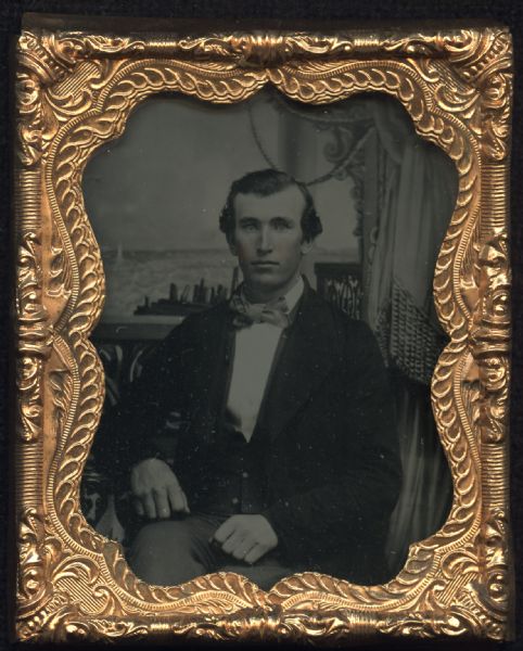 Ninth plate ambrotype of unidentified man, waist-up portrait, sitting in front of a painted land- or sea-scape backdrop, with curtains tied back on the right. He is facing forward and is wearing a suit and tie, with his elbow resting on a cloth-covered table, and his other hand in his lap. Hand-coloring on cheeks. 