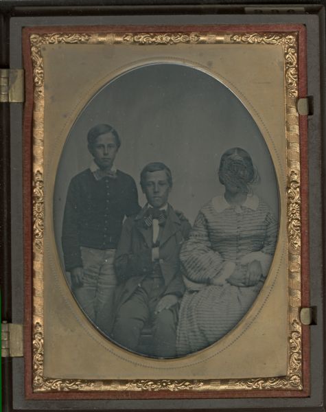 Sixth plate ambrotype of three-quarter length group portrait of a woman, sitting on the right, and two boys on the left. The boy in the center is sitting, and the boy on left is standing. All face forward.  The standing boy is wearing a boy's jacket and a tie; the boy sitting is wearing a suit and tie. The woman's face is deliberately scratched out. Hand-coloring on cheeks. 