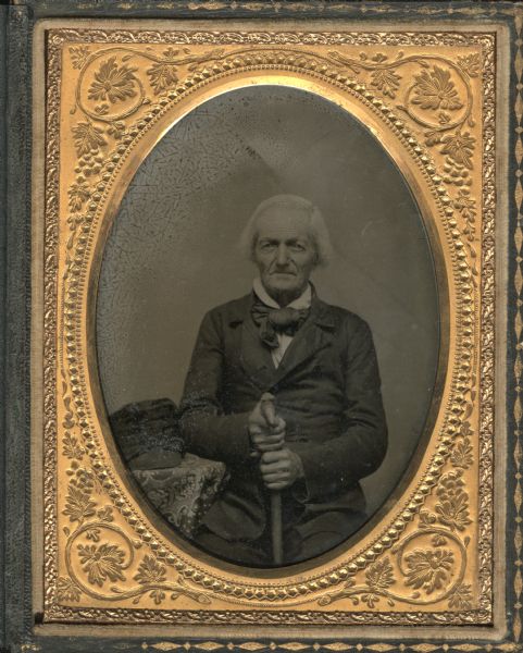 Quarter plate ambrotype of three-quarter portrait of Joseph Crelie. Seated half figure facing front with right elbow on a table, both hands holding a cane in front. He is wearing a buttoned suit coat, white shirt and large bow tie, and his hat is resting on the table. His white hair is ear length. 