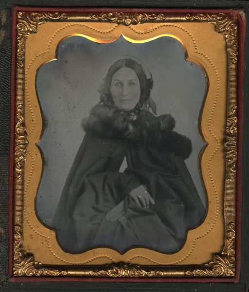 Sixth plate ambrotype of Ursula Grignon. Three-quarter length portrait, seated, facing forward, wearing a dark cape with fur wrapped around shoulders, and a bonnet tied beneath chin. Her right hand is over her left wrist, and she is wearing two rings. Hand-coloring on cheeks and rings. 