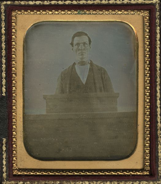 Sixth plate daguerreotype of Jedidiah K. Wood, from Oconomowoc, Wisconsin, in an unusual pose standing behind a lectern. He wears a suit and tie with the collar turned down over it. 