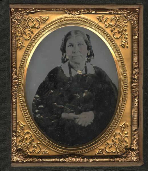 Sixth plate ambrotype of Matilda Hood, first female settler in Mineral Point, Wisconsin, in 1829. Three-quarter length portrait, seated, facing forward, hands clasped at her waist. She is wearing a dark dress, white collar with brooch at neck, and hair ribbons with drops. Hand-coloring on cheeks, earrings, and brooch. 