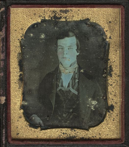Sixth plate daguerreotype of John H. Knapp. He is seated, facing forward, and is wearing a suit with velvet collar, plaid vest, cravat, and stand-up collar. His hair is combed forward over temples. Hand-coloring on cheeks and lips.