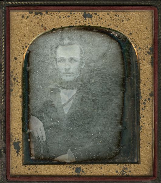 Quarter plate daguerreotype of unidentified man, a friend of Charles S. Benton of La Crosse, Wisconsin. Half length seated figure facing forward with right hand resting on chair back, wearing suit, tie, and stand collar. Hair appears to be in men's top knot style.