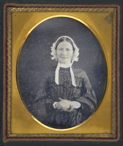 Sixth plate daguerreotype of Martha Bottomley, facing forward with hands folded at her waist. She is wearing a polka-dotted dress with pleated bodice, white collar and cuffs, and untied white bonnet. Hand-coloring on cheeks. 