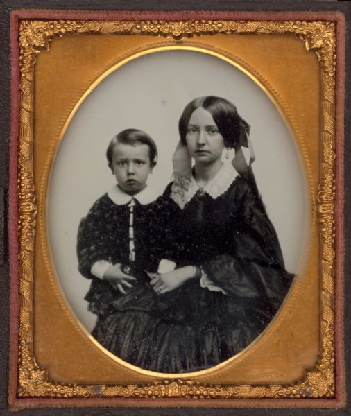 Sixth plate ambrotype of a young woman with male child. Waist-up portrait with boy standing to her right, wearing jacket and white collar and cuffs. She is wearing a dark dress, white collar, brooch at the neck, very wide, fringed hair ribbons, drop earrings, rings, and bracelet. She is holding the child's hand. Hand-coloring on cheeks. 