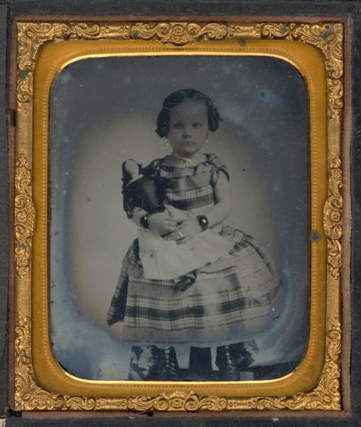 Sixth plate ambrotype of an unidentified girl with her doll. She is seated, facing front, and is wearing a plaid dress with short sleeves, drop earrings, necklace, ring, and a bracelet on each wrist. Hand-coloring on cheeks and jewelry. 