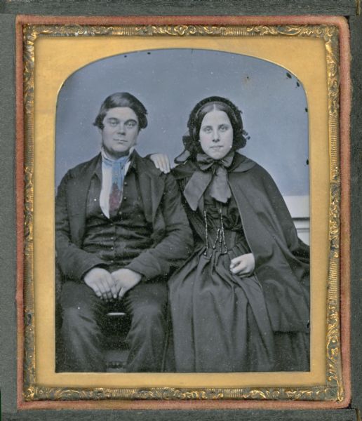 Sixth plate ambrotype of an unidentified man and woman, seated, facing front, with the woman's hand on the man's shoulder. Both are dressed in dark colors. The woman is wearing a long cape and dark bonnet tied in a large bow. The man's long tie is tinted blue and red. The backdrop is blue. Hand-coloring on cheeks. 