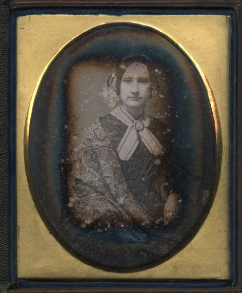 Quarter plate daguerreotype of Lucy B. Blair, seated, facing forward. She is wearing a fan front dress, lace collar, striped ribbon tied at the neck, and printed shawl draped over her shoulder. Hand-colored gold details on hair ornament.