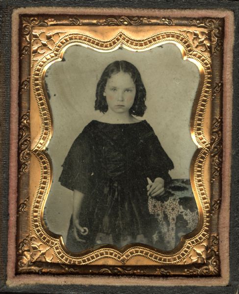 Ninth plate ferrotype/tintype of Martha Wood Crosby. Three-quarter length portrait, standing, facing forward, with left forearm on cloth covered table. She wears a 3/4-sleeve dress of fine fabric, and her right hand is holding an unidentified object. Her hair is in ringlets. Hand-coloring on cheeks and lips. 