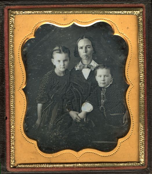 Sixth plate daguerreotype of a woman with two children. Three-quarter length portrait. The woman is seated, with her left arm around the boy, and is wearing a shawl over her dress, a cutwork white collar, checked bow tie and a choker of beads. The boy is standing next to her and is wearing a buttoned jacket over a white shirt, with his hand held still by the woman. The girl is stands on her right, leaning into her, and is wearing a printed long jacket over her dress. 