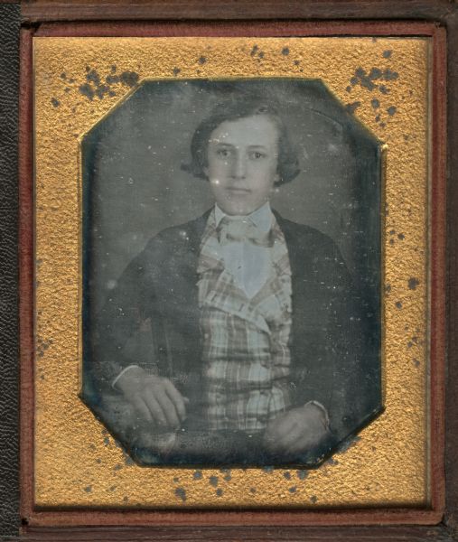 Sixth plate daguerreotype of Frank W. Burrows, brother of George B. Burrows. Waist-up portrait, facing front, with right arm resting on a table. He is wearing a suit with plaid vest and collar turned down over checked tie. Hand-coloring on cheeks.  