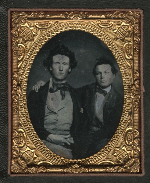 Ninth plate ambrotype of William H. Church with his schoolteacher, both sitting and facing forward. The man on the right has his arm around the shoulder of the other, and the two are holding hands. Hand-coloring on cheeks.