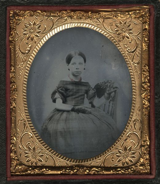 Ninth plate ferrotype/tintype of Annah Smith, daughter of John J. Smith. She is seated sideways on a chair, facing forward, with her left hand on the chair back and her right hand in her lap. She is dressed in a plaid off-the-shoulder short sleeved dress, and is wearing mitts and a red necklace. Hand-coloring on cheeks. 