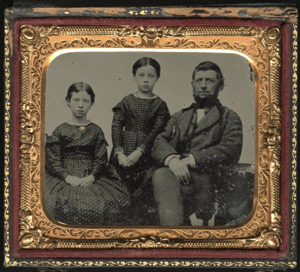 Sixth plate ferrotype/tintype of John Jay Smith with Annah and Clara R., possibly his daughters. He and the girl on the far left are sitting, and the other girl is standing between them. The girls are wearing print dresses and jewelry, and John is wearing a suit with velvet collar. Hand-coloring on cheeks and gold details on collar pin. 