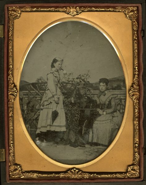 Half plate ferrotype/tintype of Clara R. Smith and G(enie?) McKnight (Ripon?). The women are dressed for the outdoors, posing amid garden props including a trellis and potted plants, in front of a painted landscape backdrop. 