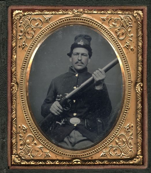 Sixth plate ferrotype/tintype of John O. Wraalstad, Sergeant, Company I. Waist-up portrait, in uniform. He is holding his rifle pointed towards the upper right. Hand-coloring on cheeks and gold buttons.