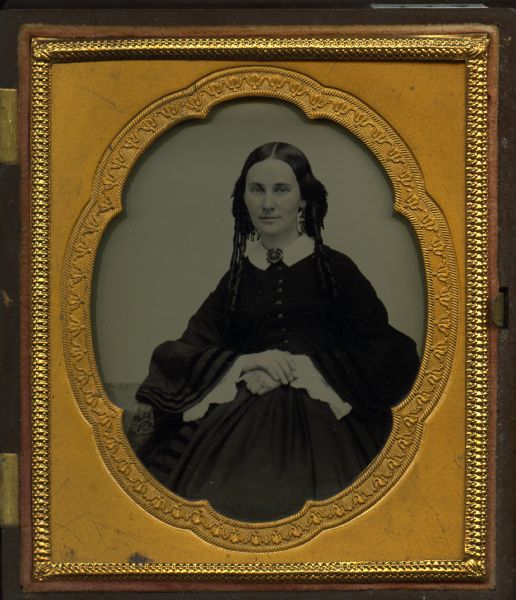 Sixth plate ambrotype of Mrs. William H. Bowman, seated next to a cloth covered table, wearing a dark dress, white collar, lace cuffs, and brooch at the neck. She has her hair in very long ringlets and adorned with lace. Hand-coloring on cheeks and lips. 