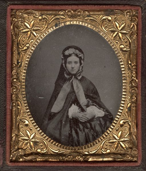 Sixth plate ferrotype/tintype of Celia Gaston. She is sitting facing forward, with hands in her lap. She is dressed for the outdoors with cape, gloves, and bonnet tied under her chin with wide ribbons and lining her face with flowers. Underneath is a dress of fine fabric. Hand-coloring on cheeks. 