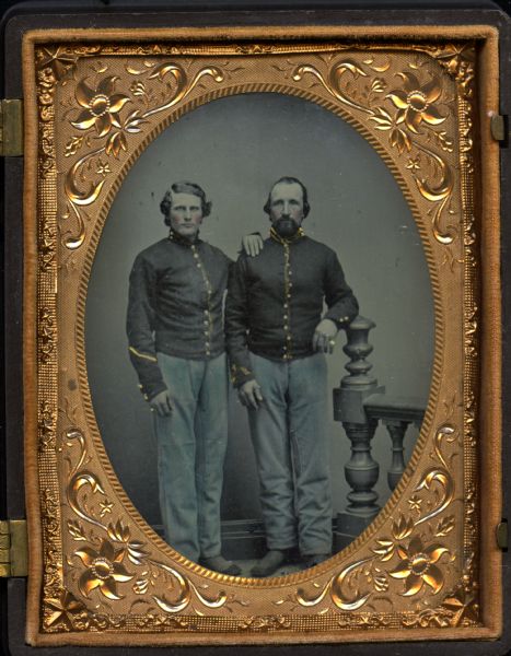 Quarter plate ferrotype/tintype. Full-length portrait of Chester Hamilton Burgess standing with an unidentified soldier. Both men are wearing Civil War uniforms with cavalry shell jackets. Burgess, on the right, has his left elbow resting on the newel post of a prop balustrade. The other soldier is resting his left hand on Burgess' shoulder. Both men are wearing little finger rings. Hand-coloring on trousers, cheeks, and gold details on uniforms and jewelry. 