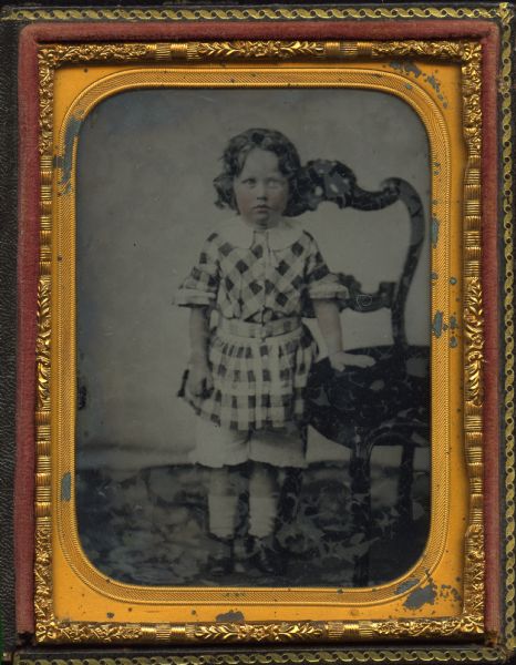 Quarter plate ambrotype of unidentified child associated with the Billinghurst family. Full-length standing portrait, wearing a belted plaid smock with white collar over knee-length white pantalettes, with hand resting on the seat of an ornate balloon back chair. Hand-coloring on cheeks and lips. 