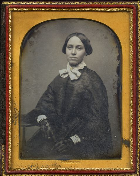 Half plate daguerreotype of Carolina Maria Field Knapp (Mrs. John H. Knapp), seated, facing forward, with her forearm resting on a table on the left. She is wearing a white collar with brooch, dark moiré cape, and black lace mitts, with a ring on her index finger. Hand-colored gold details on brooch. 