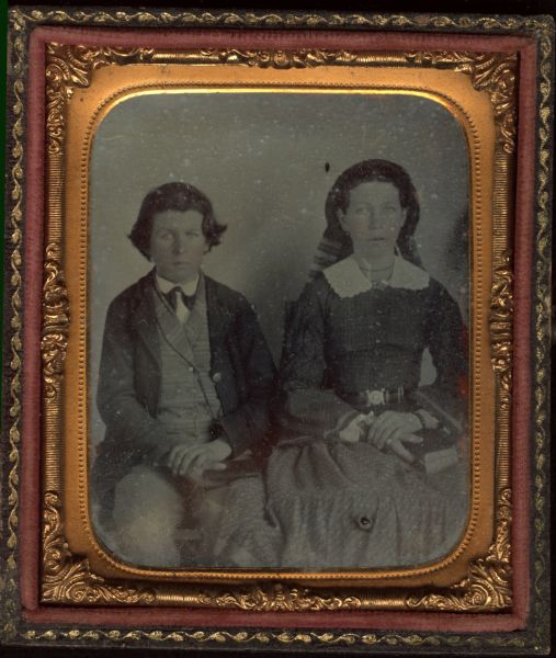 Sixth plate ambrotype of unidentified boy and girl, seated side by side, facing forward, with hands folded in laps. He is wearing a suit with striped vest, and collar folded down over his tie. She is wearing a skirt and plaid blouse, belted at the waist, with white cut-work collar, and a bonnet with plaid ribbons down the back. Each child is holding a book. Jewelry includes a bracelet, ring, and necklace. Hand-coloring on the cheeks. 