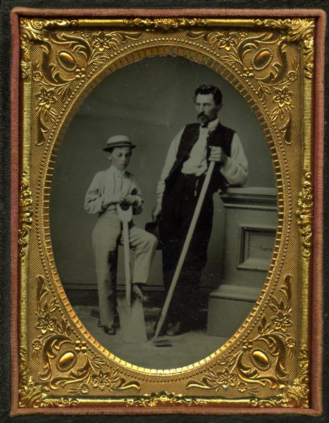 Quarter plate ferrotype/tintype of Ancil Rich and his nephew Lamont Brooks Rich. Full-length standing portrait, posed with farming tools. Ancil is holding a hoe and Lamont has a spade. Ancil leans against a studio prop balustrade and is wearing trousers, vest and shirt, and is holding his hat. Lamont is wearing a shirt, trousers, and hat, and is leaning his left foot on the spade. Hand-coloring on the cheeks. 