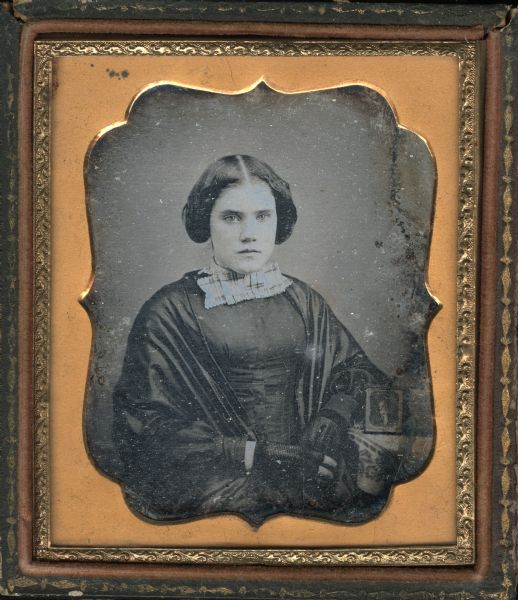 Sixth plate daguerreotype of an unidentified woman. Waist-up portrait of her seated, facing forward, with her arm resting on a cloth covered table on the right. A cased photograph is standing open on the table. She is wearing a black dress, black shawl, black fingerless gloves, and a plaid tie at the neck. 