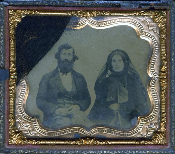Sixth plate ferrotype/tintype of a waist-up portrait of an unidentified man and woman. They are seated, facing forward, with their hands in their laps. He is wearing a jacket and double breasted striped vest with lapels. She is wearing outerwear, and a bonnet with wide ribbon ties and attached black veil down the back. 