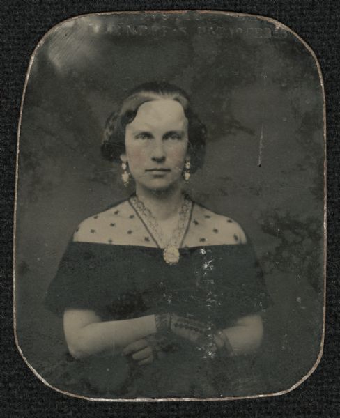 Ninth plate ferrotype/tintype of Abby O. Briggs. Waist-up portrait, facing forward. She has her arms folded across her chest, and is wearing an off-the-shoulder dark-colored gown with lace over the shoulders, dark lace fingerless gloves, drop earrings, and a brooch at the V-neck of the dress. Hand-coloring on the cheeks. 