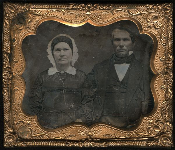 Sixth plate daguerreotype of an unidentified man and woman. Waist-up portrait, facing forward. The woman is wearing a dark dress of fine fabric, watch key chain, cutwork collar, brooch at the neck, necklace, and white cap. The man is wearing a suit and tie, with only the right side of his stand collar visible. Hand-coloring on cheeks. 