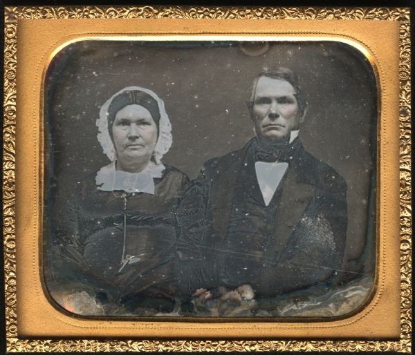 Sixth plate daguerreotype of an unidentified couple. Waist-up portrait, facing forward. The woman is wearing a dark dress of fine fabric, watch key chain, cut-work collar, brooch at the neck, necklace, and white cap. The man is wearing a suit and tie, with stand collar. Hand-coloring on cheeks. 