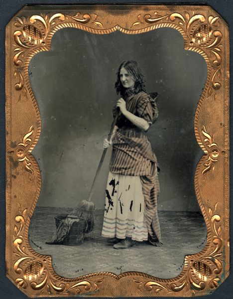 Sixth plate ferrotype/tintype full-length portrait of an unidentified young woman standing, facing left, holding a broom partially inside a bucket draped with a rag. She is dressed in a striped smock folded up and tied in the back, her straw bonnet is hanging down her back, and a kerchief is tied around her neck, and she is wearing a choker. Her tangled hair hangs loose. Hand-colored stripes on clothing. 