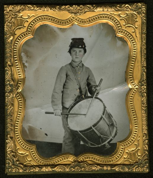 Sixth plate ferrotype/tintype of Henry A. Cooper as a drummer boy in the Union Army. Three-quarter length portrait of him standing in front of a backdrop, wearing a military uniform, and holding drumsticks over a drum slung over his shoulder. Hand-coloring on cheeks and gold details on buttons.