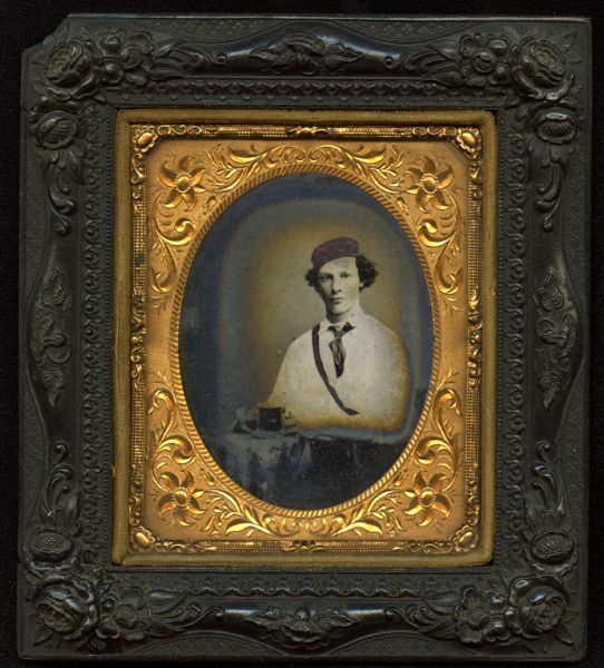 Sixth plate ferrotype/tintype of Phillip Greenthal. Waist-up portrait, seated, facing forward, in a white shirt, short tie, and cap. He is wearing a strap across his chest to carry an unseen object, and is holding, with both hands, an unidentified object on the cloth-covered table next to him. Hand-coloring on cheeks.