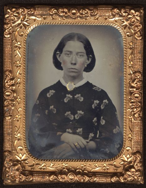 Ninth plate ferrotype/tintype of Susan Hotchkiss Searles, wife of George Searles. Waist-up portrait, facing forward. She is wearing a dark dress with a lighter flower print, a small white cutwork collar and brooch at the neckline. Her hands are folded at her waist. Hand-coloring on her cheeks and lips, and gold details on brooch, earring, and ring. 