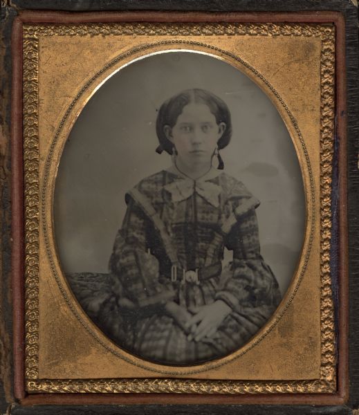 Sixth plate ambrotype of Susan Hotchkiss Searles, wife of George Searles. She is seated next to a cloth-covered table, facing forward with hands in lap, wearing a belted fan-front plaid dress, drop earrings, choker holding a large bow, and ribbons in her hair. Hand-coloring on cheeks. 