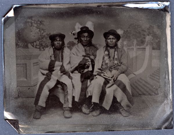 Sixth plate ferrotype/tintype of three Woodland Native Americans. Full-length group portrait in front of a painted classical garden studio backdrop. They are seated, facing forward, each wearing a hat and moccasins, and wrapped in Hudson Bay blankets. The man in the center appears older than the other two, and has feathers in his hat. The younger men have long braids, and the man on the right is wearing a beaded shirt. 