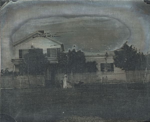 Quarter plate ambrotype of a white house with dark shutters, probably located in Madison, Wisconsin. The house has a gable end at the left, with a portico off of the first floor, and and a one-story addition to the right. Three small trees and a white picket fence obscure parts of the facade. A woman and child are standing in front of the picket fence. She is holding a pram with another child sitting in it. 