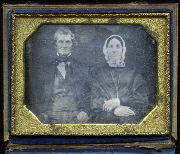 Quarter plate daguerreotype of Mr. and Mrs. Oakley, parents of Mrs. Sylvanus Wade of Greenbush, Wisconsin. Waist-up portrait, seated, facing forward, with hands in laps. Mr. Oakley is wearing plaid trousers, suit, cravat, stand collar, and watch key chain. Mrs. Oakley is wearing a dark dress, lace collar, tied white bonnet, gold chain necklace, and spectacles. 